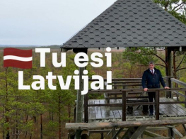 Skatu tornī stāv cilvēks tumšās drēbēs, redzams grafisks Latvijas karogs un baltiem burtiem uzraksts "Tu esi Latvija!"