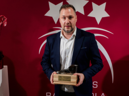 Vīrietis uzvalkā ar apbalvojumu rokās stāv pie sarkanas sienas ar baltām zvaigznēm