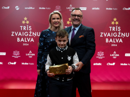 Sieviete zilā kleitā  un blondiem matiem kopā ar vīrieti uzvalkā un bērnu, kurš tur rokās zelta ķiegeļi, trofeju.