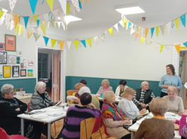 Seniori sēž istabā pie galdiņiem un apglezno baltus traukus