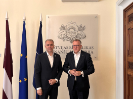 Divi vīrieši melnos uzvalkos stāv telpā pie valstu karogiem