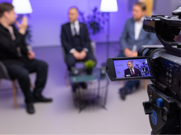 trīs vīrieši tumšās drēbēs sēž krēslos un priekšplānā filmēšanas kamera