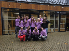 Jauniešu grupas kopbilde krāsainos tērpos pie brūnas ēkas