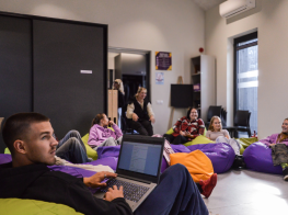 Jaunieši sēž uz krāsainiem pufiem ar datoriem klēpī gaišā telpā