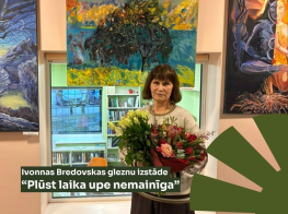 gleznotāja ar ziediem rokās un fonā viņas gleznas